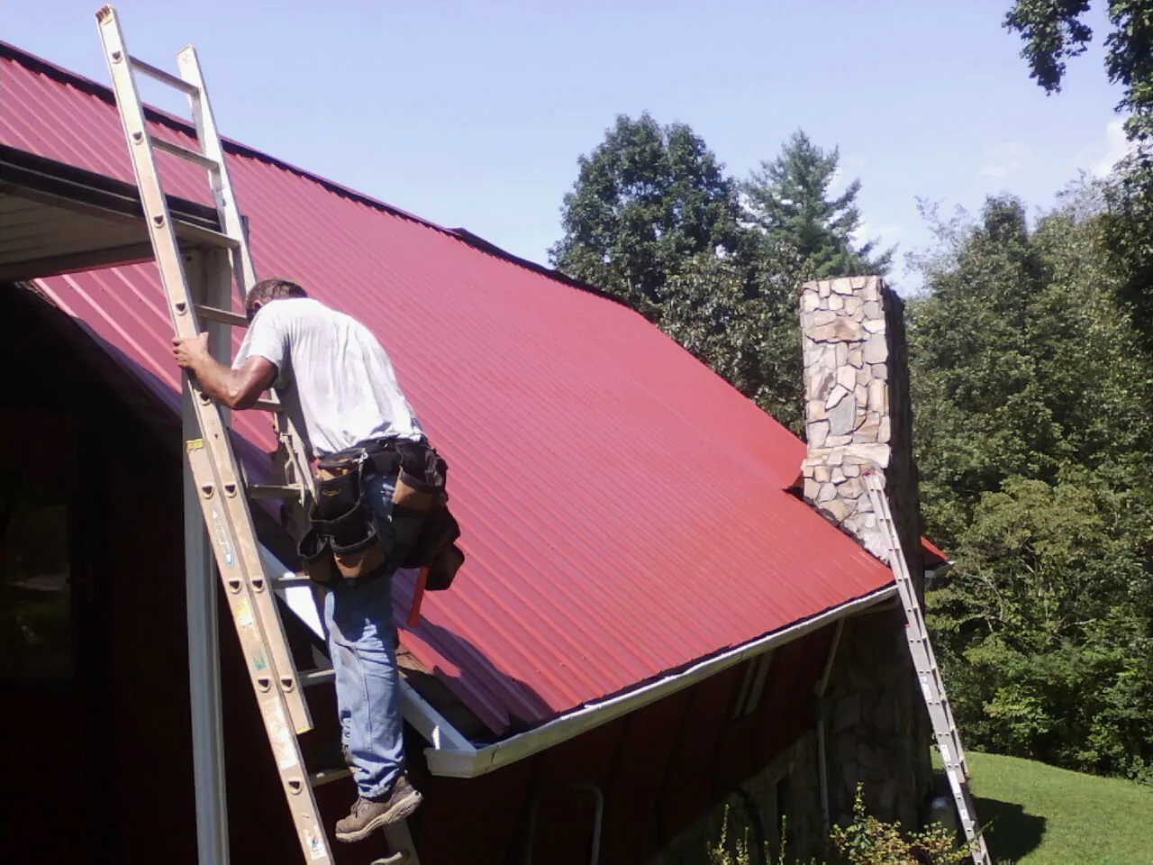 Roofer on a ladder beside a red metal roof and stone chimney — Soffit & Fascia Repair by Roofing Brothers in Monona